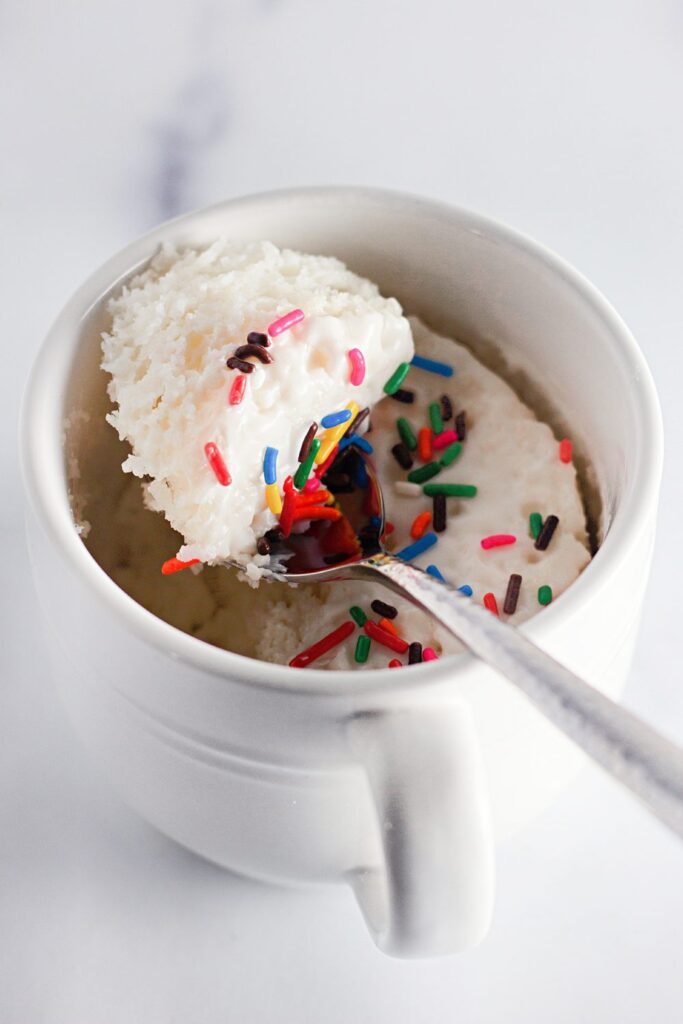 Finished white cake in a mug with vanilla frosting and sprinkles with a spoon dipped into the cup